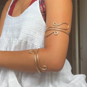 Puede incluir: Dos brazaletes de metal en tono dorado con un diseño de alambre liso y continuo con extremos curvados. Un brazalete se coloca en la parte superior del brazo, mientras que el otro se lleva en el antebrazo, ambos con una estructura metálica curva.