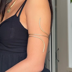 Könnte beinhalten: Ein Oberarmreif aus goldfarbenem Metall mit einem glatten, spiralförmigen Drahtdesign, das sich um den Oberarm legt. Das Schmuckstück besteht aus einem durchgehenden, schmalen Band, das an den Enden nach oben und unten gebogen ist und eine offene Form bildet.