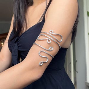May include: A silver-toned metal arm cuff featuring a continuous wire design that wraps around the upper arm. The accessory is constructed from a single piece of smooth, polished wire formed into a series of rounded, flowing, and interconnected loops.
