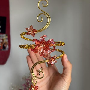May include: A gold-colored wire arm cuff featuring a spiral design adorned with several translucent pink flower-shaped accents. The wire is shaped into elegant curves and swirls, with the pink floral elements attached at various points along the metal.