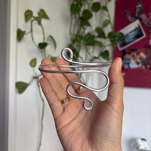 Könnte beinhalten: Ein Oberarmreif aus silberfarbenem Metall mit einem durchgehenden Drahtdesign, das sich an den Enden zu zwei markanten, abgerundeten Schlaufen biegt. Das Schmuckstück besteht aus einem einzelnen, glatten, polierten Draht in offener Form.
