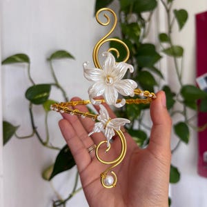 May include: A gold-toned wire arm cuff featuring a large white lily flower and a smaller white butterfly accent. The wire is shaped into elegant swirls and is embellished with small white pearls along the band and at the terminal ends.
