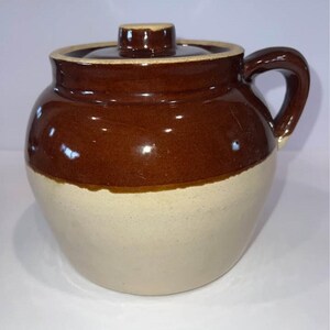 Op de afbeelding: Een ronde keramische pot met een deksel en een enkel handvat aan de zijkant. Het bovenste gedeelte van het vat is afgewerkt met een glanzend donkerbruin glazuur, terwijl het onderste gedeelte een matte, lichtbeige afwerking heeft.