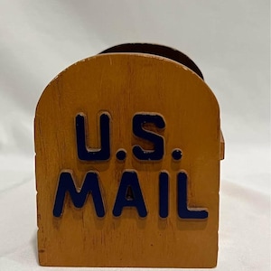 May include: A wooden desk organizer shaped like a classic mailbox with a rounded top. The front panel is finished in a light brown wood tone and features the text U.S. MAIL in raised, dark blue capital letters centered on the surface.