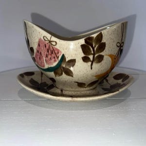May include: A speckled ceramic gravy boat and matching saucer set featuring a hand-painted pattern of pink watermelon slices, dark brown leaves, and orange accents. The gravy boat has a distinctively curved, asymmetrical rim and a wide spout.
