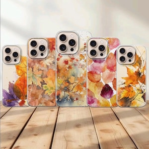 Puede incluir: Cinco fundas transparentes para smartphone con varios patrones de hojas de otoño estilo acuarela en tonos naranja, amarillo, rojo y morado. Cada funda tiene un recorte para un sistema de triple cámara y están alineadas sobre madera.
