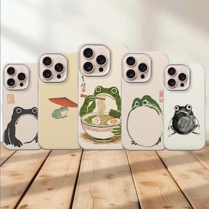 May include: Five smartphone cases featuring various frog illustrations in an ink-wash style. Designs include a frog with a red umbrella, a frog eating ramen with chopsticks, a circular frog outline, and two distinct frog portraits with red stamps.