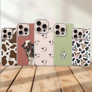 Op de afbeelding: Vijf smartphonehoesjes met koe-thema. De eerste heeft een bruin-witte koeienprint. De tweede is oudroze met een koeienportret. De derde is lichtroze met kleine koeiengezichtjes. De vierde is saliegroen met een kleine koe die ballonnen vasthoudt. De vijfde heeft een zwart, wit en bruin koeienpatroon.