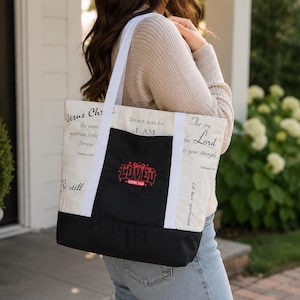 Può includere: Una borsa tote in tela color crema con spallacci bianchi e un pannello inferiore nero. La borsa presenta una tasca centrale nera con la parola LOVED ricamata in rosso e testo che include Jesus Christ, Do not fear e The joy of the Lord.