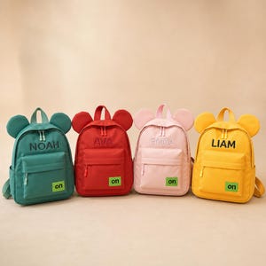 Peut inclure: Quatre sacs à dos en bleu sarcelle, rouge, rose clair et jaune, chacun avec des oreilles arrondies sur le dessus, une poche avant zippée et un petit écusson rectangulaire vert avec le texte 'on'. Les noms 'NOAH', 'AVA', 'EMMA' et 'LIAM' sont brodés.