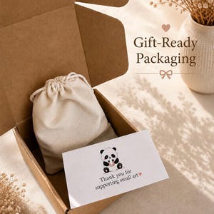 May include: A brown cardboard shipping box containing a cream-colored drawstring pouch and a white rectangular card. The card features a black and white panda illustration and the text: Thank you for supporting small art. The text Gift-Ready Packaging is visible.