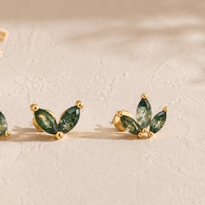 May include: Three gold stud earrings featuring green moss agate stones. One earring has a single pear-shaped stone, one has two marquise-shaped stones, and one has three marquise-shaped stones arranged in a fan-like cluster with gold metal settings.