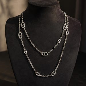 Könnte beinhalten: Eine lange silberne Halskette mit gleichmäßig verteilten ovalen Gliedern, die jeweils einen horizontalen Steg in der Mitte aufweisen. Die Kette ist zweireihig drapiert und zeigt eine polierte metallische Oberfläche sowie ein klassisches Design.