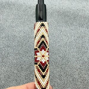 May include: A black ink pen featuring a cylindrical barrel covered in a hand-beaded sleeve. The beadwork displays a geometric pattern in a repeating diamond motif, utilizing a color palette of cream, deep red, black, and metallic gold glass beads.