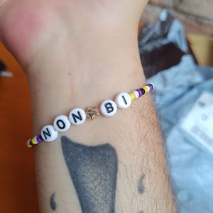 Pode incluir: Uma pulseira de contas com pequenas sementes amarelas, brancas, roxas e pretas. A pulseira inclui contas redondas brancas com letras pretas que soletram NON BI, separadas por uma pequena conta metálica em forma de estrela no centro.