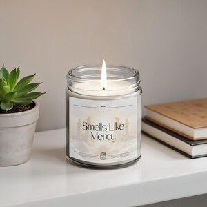Peut inclure: Une bougie en pot de verre transparent avec une mèche allumée, dotée d'une étiquette blanche affichant une croix, le texte "Smells Like Mercy" et une illustration d'un escalier menant à des piliers dans les nuages. La cire est blanche.