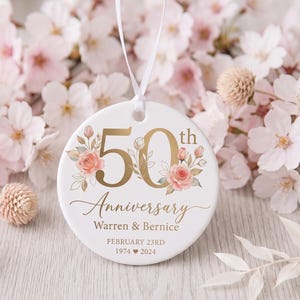 Puede incluir: Un adorno redondo de cerámica blanca con texto dorado que dice 50th Anniversary, Warren & Bernice, February 23rd, 1974 corazón 2024. El diseño incluye ilustraciones de rosas rosadas y hojas verdes, y cuelga de una cinta blanca fina.