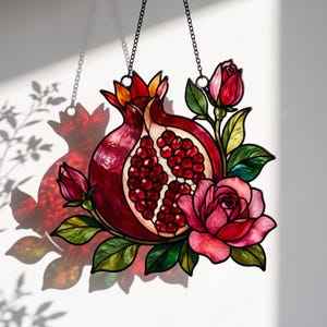 Peut inclure: Un attrape-soleil en vitrail représentant une grenade coupée en deux avec des graines rouge foncé, entourée de roses roses et de feuilles vertes. La pièce est suspendue par une fine chaîne métallique, révélant des couleurs vives.