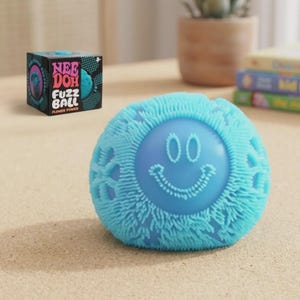 May include: A light blue Nee Doh Fuzz Ball with a textured, fuzzy surface and a smooth center featuring a raised smiley face design. The ball is accompanied by its black square packaging box, which displays the text Nee Doh Fuzz Ball Flower Power.