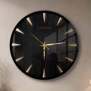 Puede incluir: Un reloj de pared circular con una esfera negra que presenta una sutil textura de veta de madera, marcadores de hora triangulares dorados y manecillas doradas finas. El texto "CALM+CERO" está impreso en dorado cerca de la parte superior.