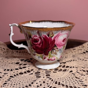 May include: A white porcelain teacup featuring a vibrant floral pattern of deep burgundy and soft pink roses. The cup has a scalloped rim with a gold-painted edge and a decorative, ornate handle. The base is pedestal-style with gold trim.