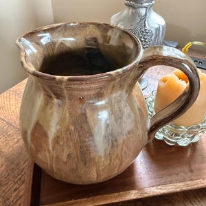 May include: A ceramic pitcher featuring a rounded body and a curved handle, finished with a reactive glaze in shades of tan, brown, and cream that creates a dripping effect. The pitcher has a wide, open mouth with a small pouring spout.