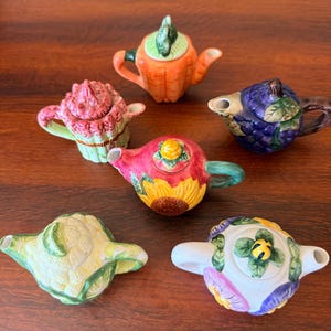 May include: A collection of six miniature ceramic teapots shaped like various fruits and vegetables, including a carrot, a cluster of grapes, a sunflower, a head of cauliflower, and a floral design, each with a matching lid and handle.