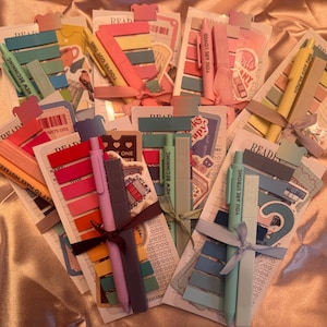 May include: A collection of stationery sets, each featuring a rectangular card with the word READING printed at the top, accompanied by a pen, a highlighter, and multiple adhesive page markers in various colours, all tied together with a ribbon.