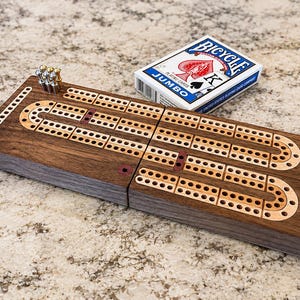 May include: A rectangular wooden cribbage board featuring a dark brown wood grain finish with inlaid light wood tracks and metal pegs. A white box of Bicycle Jumbo playing cards is positioned next to the board, displaying the brand name and logo.