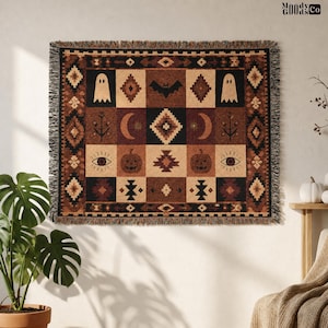 May include: A woven throw blanket featuring a grid pattern with brown, black, and cream colors. The design includes icons of ghosts, bats, pumpkins, crescent moons, and eyes, surrounded by a geometric border with fringe on the top and bottom edges.