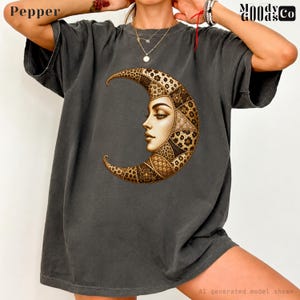 May include: A charcoal gray short-sleeve t-shirt featuring a large graphic of a crescent moon with a human face profile. The moon is decorated with intricate floral and geometric patterns in shades of gold and brown. The text Pepper and Moody Goods Co appear in the corners.