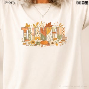 May include: An ivory t-shirt featuring the word THANKFUL in large block letters with various autumn-themed patterns including plaid, polka dots, and stripes. The design is decorated with orange pumpkins, brown mushrooms, sunflowers, and fall leaves.