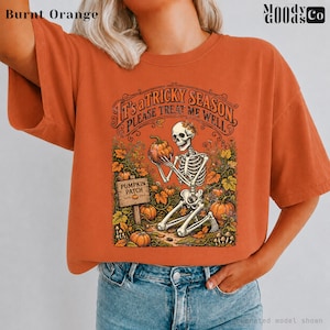 May include: A burnt orange t-shirt featuring a graphic of a skeleton kneeling in a pumpkin patch. The text reads, "It's a tricky season, please treat me well" and "Pumpkin Patch" on a wooden sign. The design includes orange pumpkins and foliage.