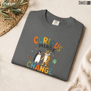 May include: A charcoal gray t-shirt featuring a graphic of a penguin wearing a bow tie and a tiger wearing a hat. The text reads CURIOUS MINDS CHANGE, accompanied by small star and globe illustrations in orange, blue, and green colors.