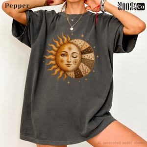May include: A charcoal gray short-sleeve t-shirt featuring a large graphic print of a sun and moon face. The sun has golden rays, while the moon side displays intricate patterned segments. Small stars are scattered around the central celestial design.