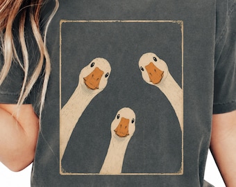 以下が含まれることがあります： チャコールグレーのクルーネックTシャツ。オレンジ色のくちばしをした3羽のベージュのガチョウが下を見下ろしている長方形のグラフィックが描かれています。左上に「Pepper」、右上に「Moody Goods Co」のロゴがプリントされています。