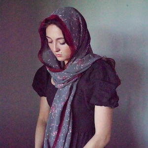 May include: A gray head covering featuring a small pink floral pattern and a deep red lace trim along the edges. The fabric is draped over the head and around the neck, with the text Angelus Rose Veils visible at the top of the frame.