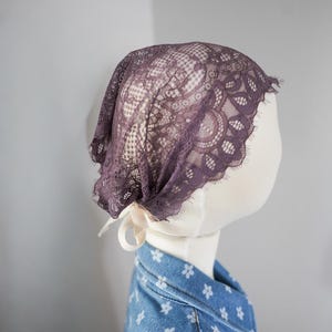 May include: A dark purple lace head covering featuring a floral pattern with scalloped edges and a delicate fringe. The lace is secured at the nape of the neck with a thin, light-colored ribbon tie, creating a soft and intricate appearance.