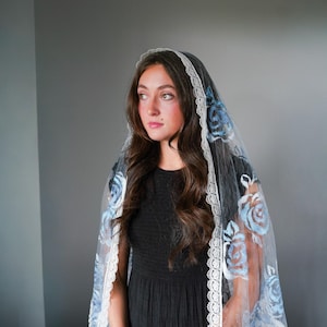 May include: A sheer chapel veil featuring a delicate white lace trim along the edges and embroidered light blue rose patterns throughout the fabric. The veil is draped over the head and shoulders, showcasing a scalloped lace border with fringe.