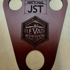 May include: A brown leather woggle featuring three circular cutouts. The surface is laser-engraved with the text NATIONAL JST and a hexagonal emblem containing the words SCOUTING AMERICA, ELEVATE, and NATIONAL JAMBOREE with a mountain graphic.