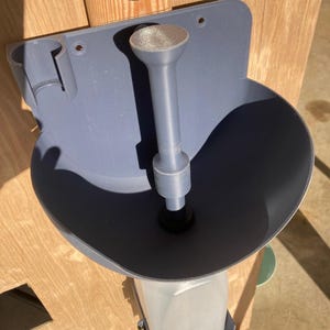 May include: A gray plastic funnel with a wide, bowl-shaped top and a long, cylindrical spout. The funnel is mounted on a wooden surface and has a small holder on the side. The design includes a clear plastic container below the spout.