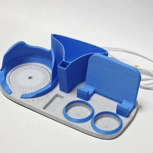 May include: A blue and light gray desk organizer featuring a circular wireless charging pad with a lightning bolt icon, a vertical phone stand, two circular pen holders, and a triangular storage compartment, accompanied by a white power cable.