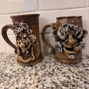 以下が含まれることがあります： 立体的な顔が彫刻された2つの茶色の陶器製マグカップ。左のマグカップはカールした髪と目立つ歯を持つ人物を描いています。右のマグカップは角、大きな鼻、顔と耳に質感のあるディテールを持つ人物を描いています。