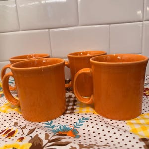 Può includere: Un set di quattro tazze in ceramica color arancione brillante con corpi lisci e arrotondati e manici curvi. Ogni tazza presenta una smaltatura arancione solida e uniforme e un bordo superiore ampio e aperto, disposte in gruppo.