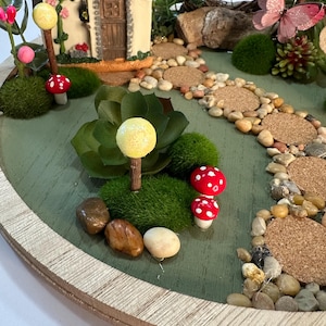 K&ouml;nnte beinhalten: Eine Miniatur-Feengarten-Szene auf einer runden Holzbasis. Sie zeigt ein kleines Haus, einen Weg aus Steinen und dekorative Elemente wie Pilze, einen Schmetterling und Gr&uuml;nzeug. Die Farbpalette umfasst Gr&uuml;n-, Braun- und Rott&ouml;ne.