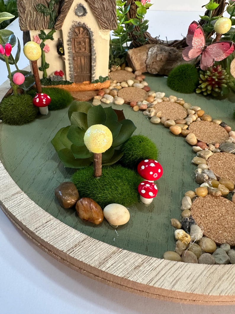K&ouml;nnte beinhalten: Eine Miniatur-Feengarten-Szene auf einer runden Holzbasis. Sie zeigt ein kleines Haus, einen Weg aus Steinen und dekorative Elemente wie Pilze, einen Schmetterling und Gr&uuml;nzeug. Die Farbpalette umfasst Gr&uuml;n-, Braun- und Rott&ouml;ne.