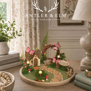 K&ouml;nnte beinhalten: Eine Miniatur-Feengarten-Szene mit einem kleinen Haus, Schmetterlingen und einem Steinweg. Die Szene befindet sich auf einem Holztablett mit gr&uuml;nem Untergrund und ist von dekorativen Elementen umgeben. Der Text "ANTLER & ELM" und "ETSY.COM" sind sichtbar.