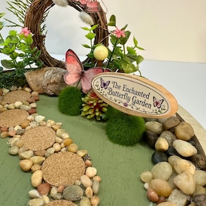 K&ouml;nnte beinhalten: Eine Miniatur-Feengarten-Szene mit einem gewundenen Steinweg und einem Schild mit der Aufschrift "The Enchanted Butterfly Garden". Die Szene umfasst einen kreisf&ouml;rmigen Zweigkranz, rosa Schmetterlinge und kleine gr&uuml;ne Pflanzen. Der Weg besteht aus kleinen Steinen und Korktrittsteinen.