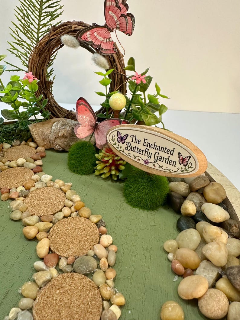 K&ouml;nnte beinhalten: Eine Miniatur-Feengarten-Szene mit einem gewundenen Steinweg und einem Schild mit der Aufschrift "The Enchanted Butterfly Garden". Die Szene umfasst einen kreisf&ouml;rmigen Zweigkranz, rosa Schmetterlinge und kleine gr&uuml;ne Pflanzen. Der Weg besteht aus kleinen Steinen und Korktrittsteinen.
