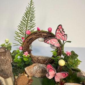 K&ouml;nnte beinhalten: Eine Miniatur-Feengarten-Szene mit einem braunen Zweigkranz mit rosa Akzenten, einem kleinen Haus und dekorativen Schmetterlingen. Die Szene umfasst k&uuml;nstliches Gr&uuml;n, kleine Steine und ein Schild mit der Aufschrift "The Enchanted Butterfly".